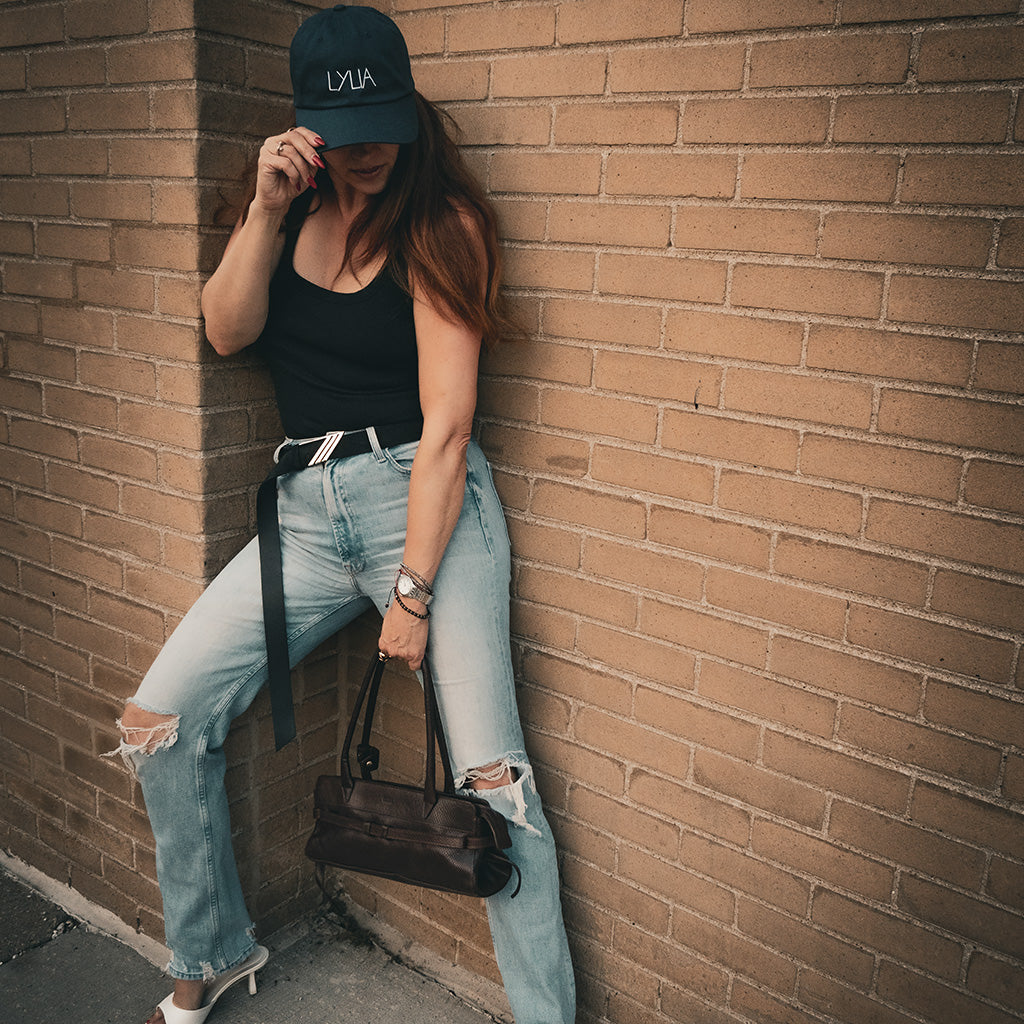 Female artist wearing a black cap and ripped jeans leaning against a brick wall.
