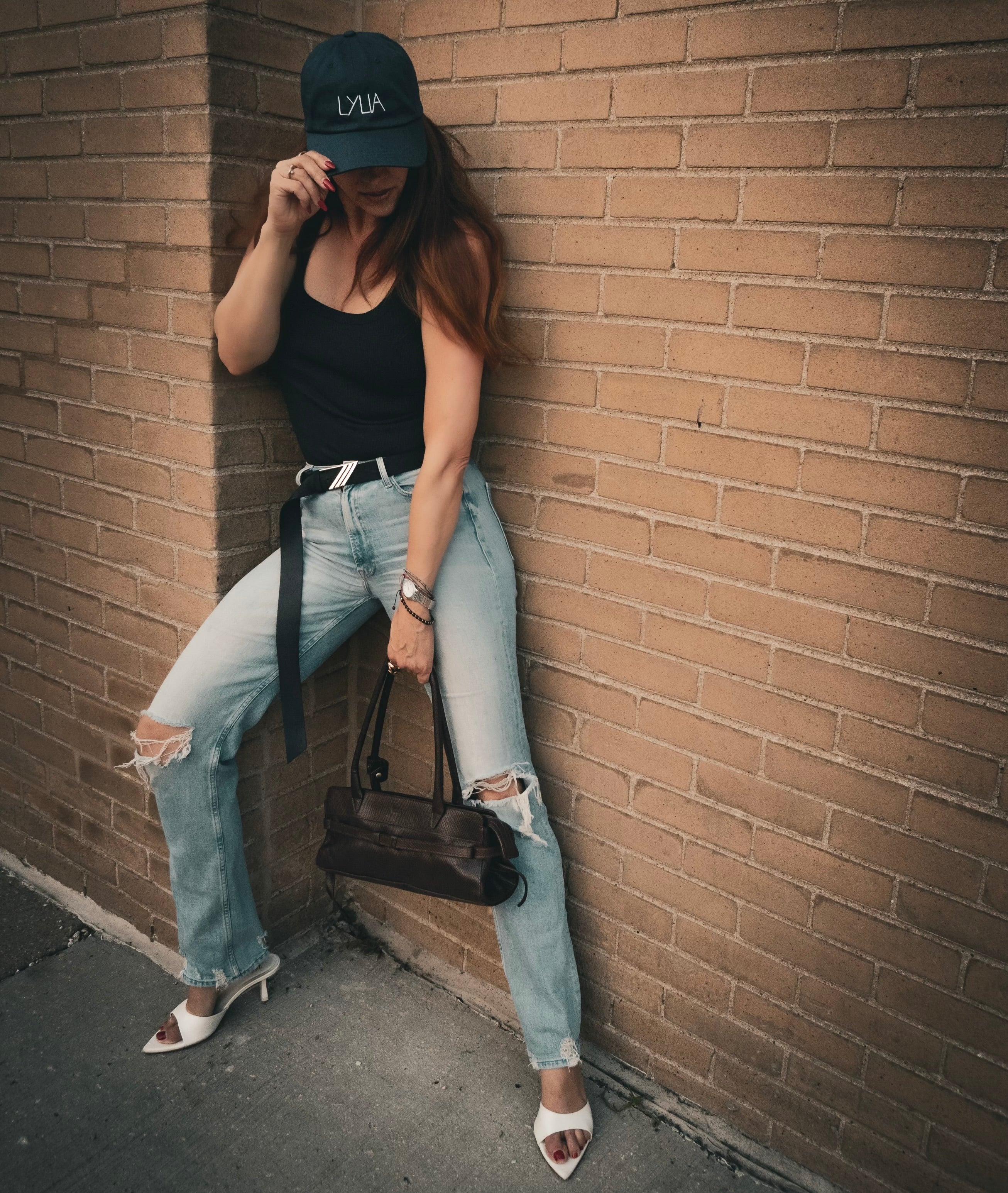 Female artist wearing a black cap, black top, and ripped jeans leaning against a brick wall.